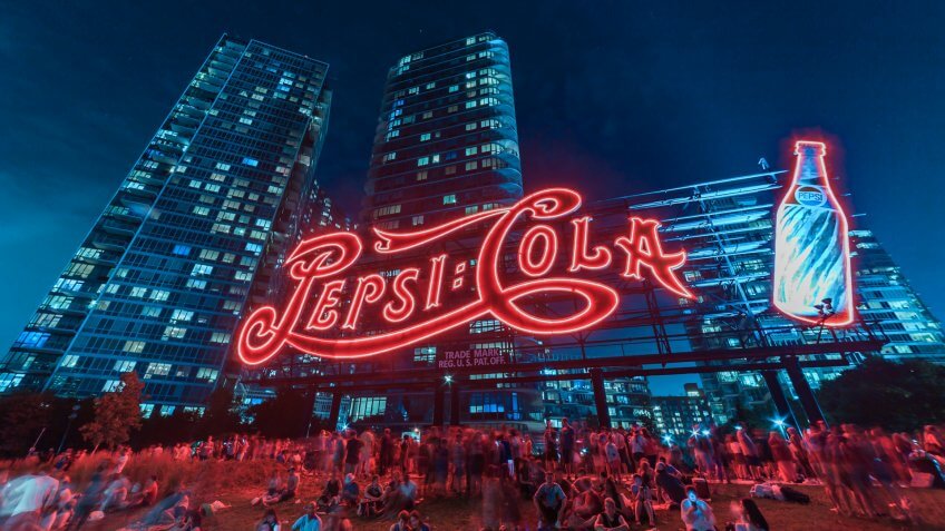 Image shows the Pepsi Cola sign and many people sitting on the ground during the celebration of the Independence day at night.