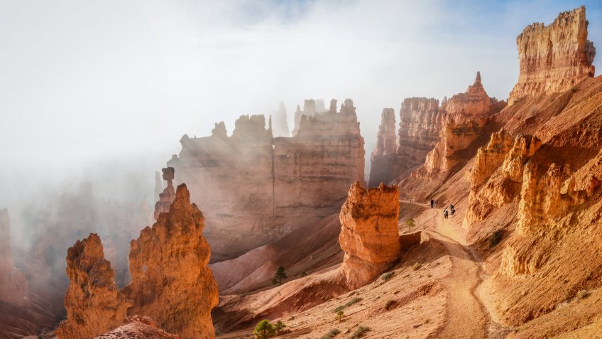 People trekking the Navajo loop trail in Bryce Canyon National Park at sunrise with some clouds and fog.