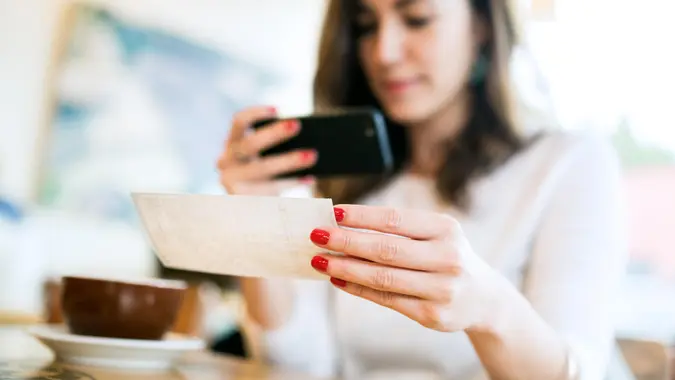 A smiling young woman takes a picture with her smart phone of a check or paycheck for digital electronic depositing, also known as "Remote Deposit Capture".