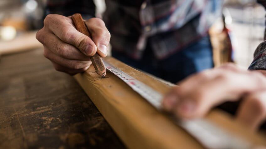 Close up of male worker using pen while drawing measurements on a wood plank in a carpentry workshop.