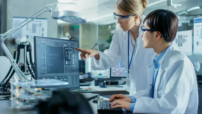 Asian Scientist Sitting at His Desk Consults Senior Engineer about Sophisticated Coding and Programming.