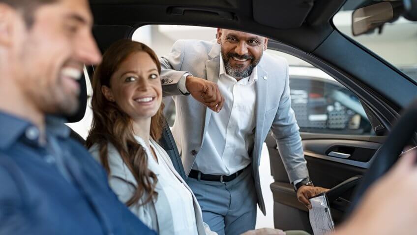 African salesman showing new car to young couple in showroom.
