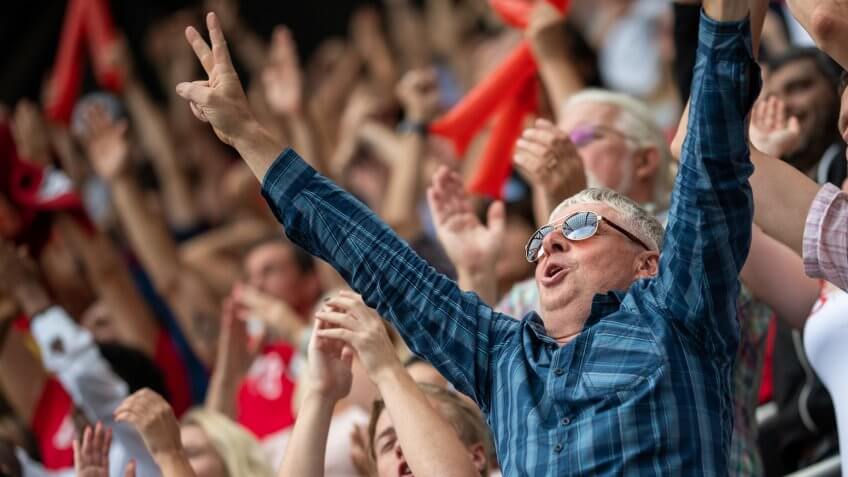 Portrait of a middle-aged man with sunglasses raising his arms in reaction to his team winning.