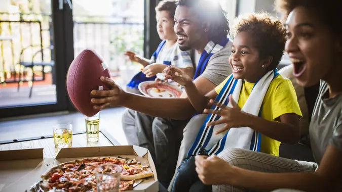 Young joyful black family having fun while cheering for their favorite American football team at home.