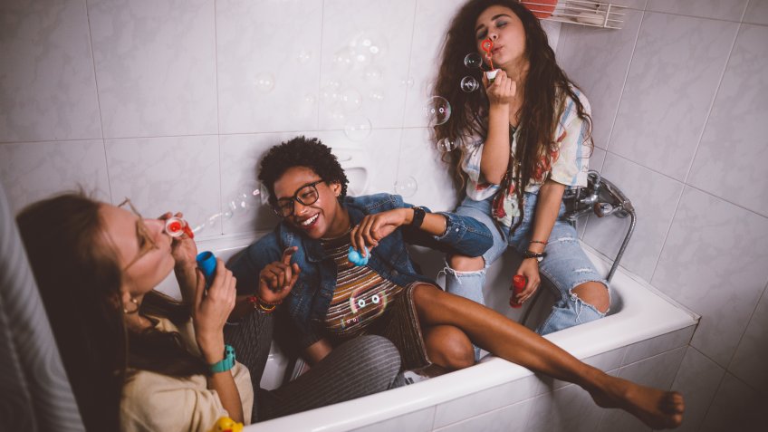 Young multi-ethnic hipster girls having slumber party at home and blowing bubbles in bathtub.