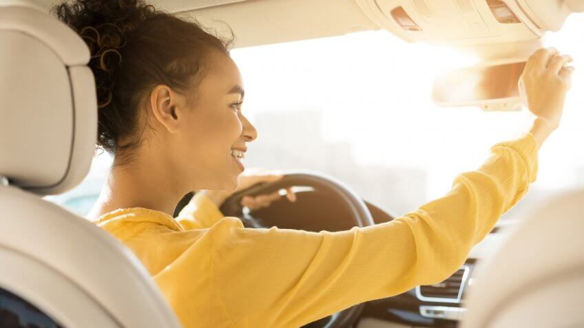 Happy black girl touching rear view mirror, driving car and smiling, copy space.