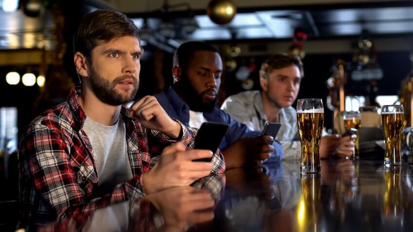 Nervous fans checking bets on smartphone, watching game in bar, bookmaker app.