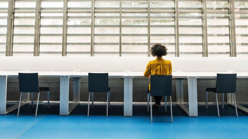 Rear view of young student or businesswoman sitting on desk in room in a library or office.