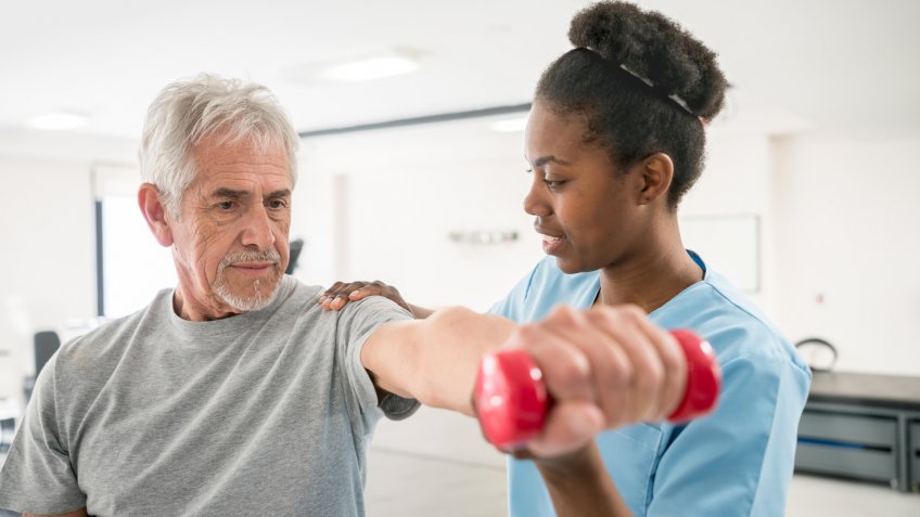 Physiotherapist correcting her senior patient with his shoulder posture as he lifts free weights both looking focused and smiling.