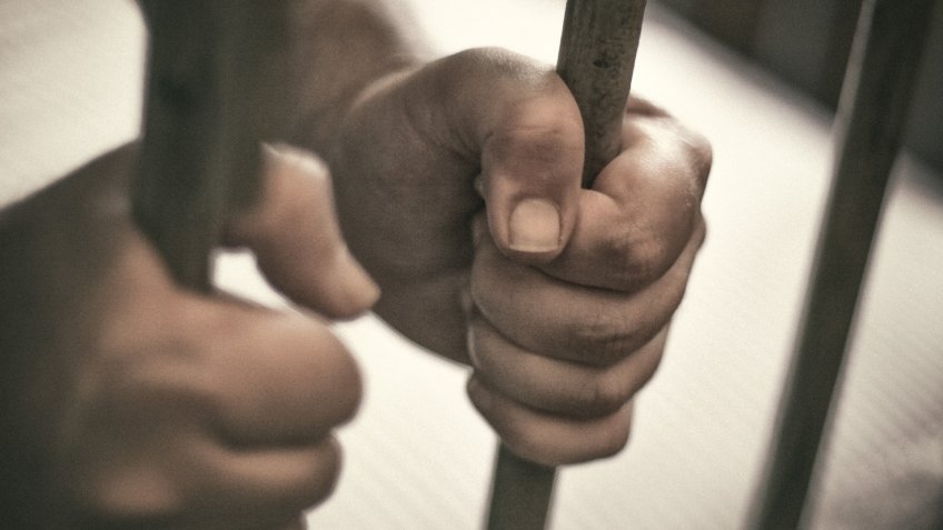Man's hands tightly holding the bars of his jail cell.