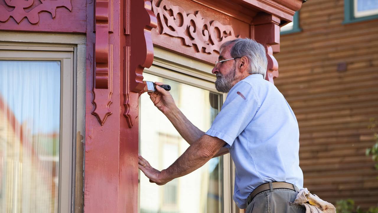 An older man in an historic town paints his house, with careful attention to detail, while standing on a ladder.