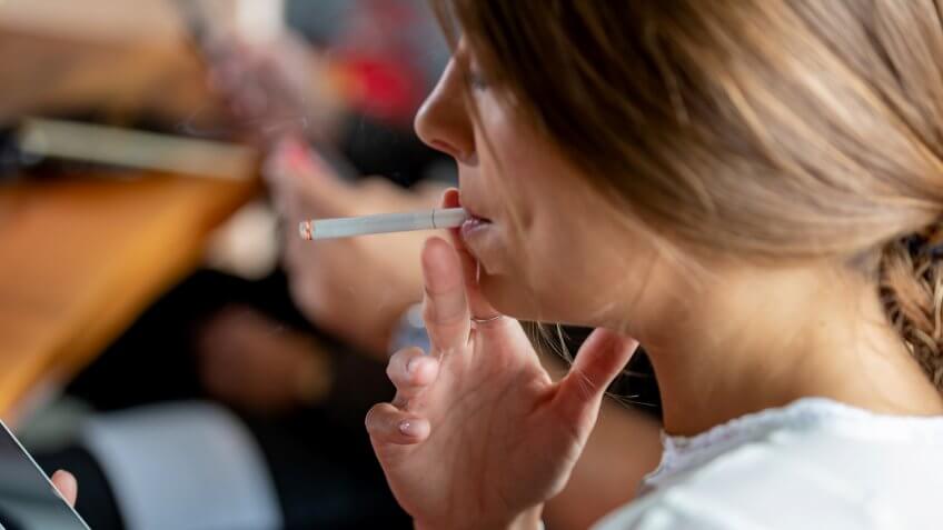 Businesswoman looking at smartphone, smoking cigarette on lunch break in sushi bar.