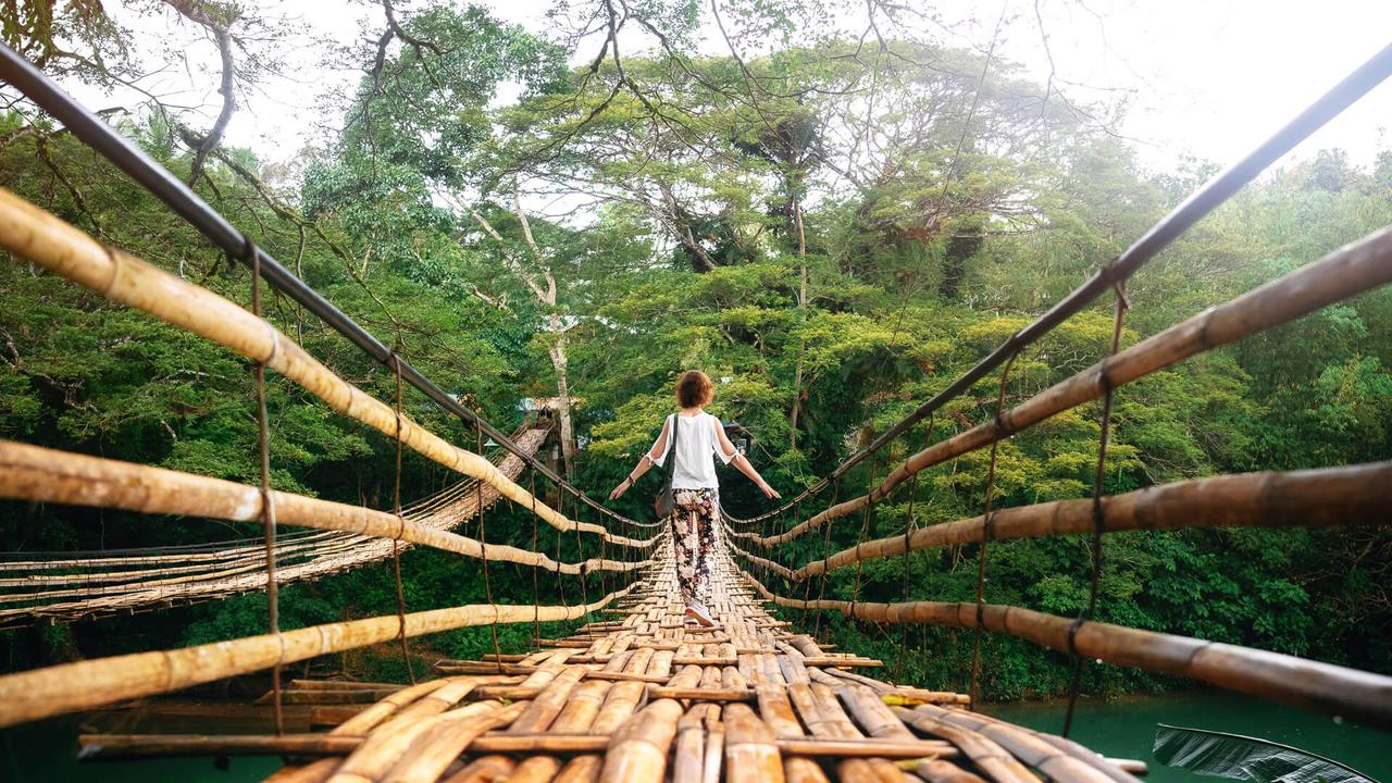 Back view of young woman on suspension wooden bamboo bridge across Loboc river in jungle.