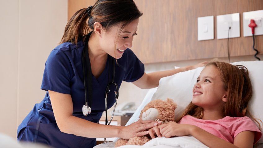 Female Nurse Visiting Girl Lying In Hospital Bed Hugging Teddy Bear.