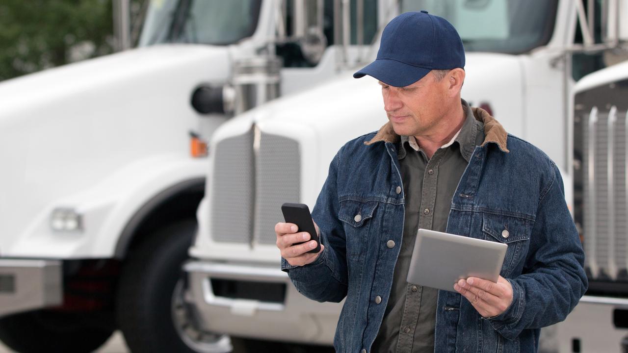 A royalty free image from the trucking industry of a truck driver using a cellphone and tablet computer in front of semi trucks.
