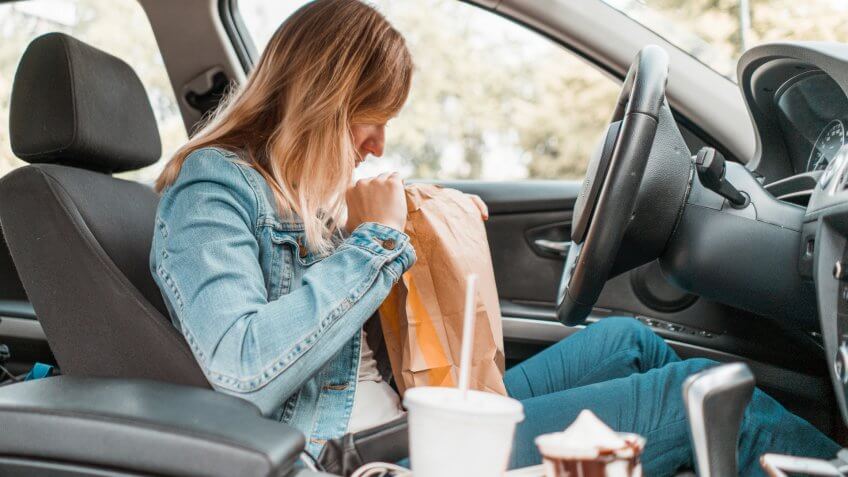 Young woman sitting in her car, opening a take out bag, eating fast food and drinking soda and coffee.