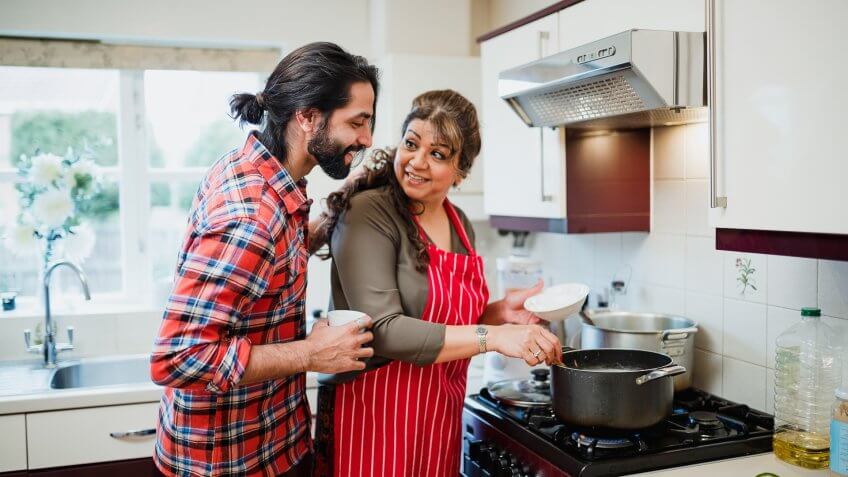 Mature woman is cooking in her kitchen and her son is looking over her shoulder to see what she is making.