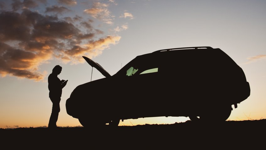 A silhouette of a woman dialing for help beside her vehicle having mechanical difficulty.