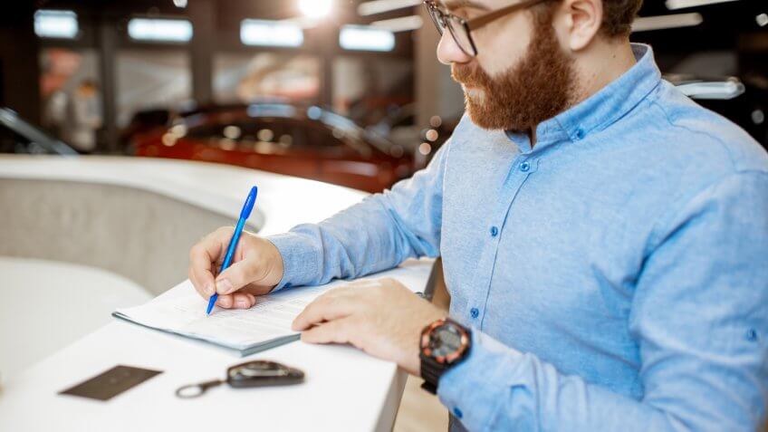 Young man signing some documents, buying or renting a car in the modern showroom of the car dealership.
