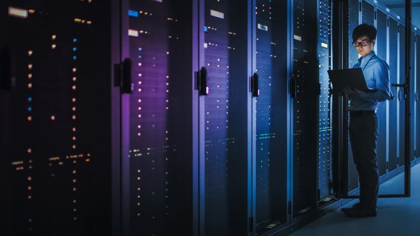 In Dark Data Center: Male IT Specialist Stands Beside the Row of Operational Server Racks, Uses Laptop for Maintenance.