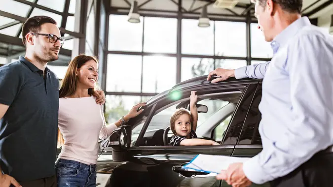 Young happy parents communicating with salesperson while buying a car in a showroom.