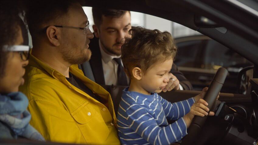 Side view of happy parents with kid sitting inside of car in auto shop and talking to consultant about purchase.