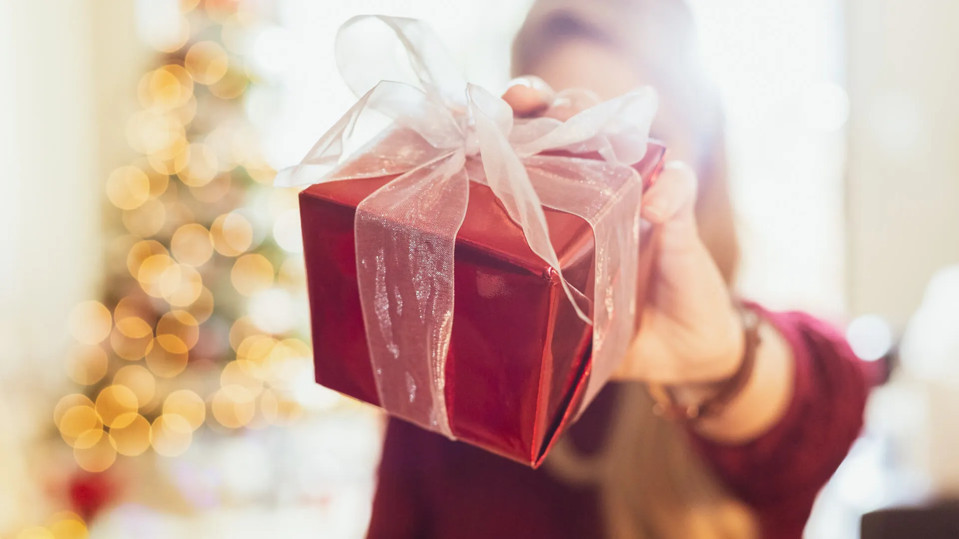 Unrecognizable woman holds a Christmas present wrapped in red paper and tied with a white transparent bow.