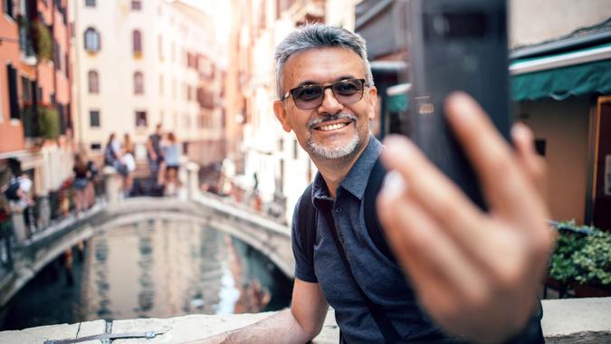 Happy handsome senior bearded man smiling while taking selfie picture with mobile phone and wearing eyeglasses outdoors.