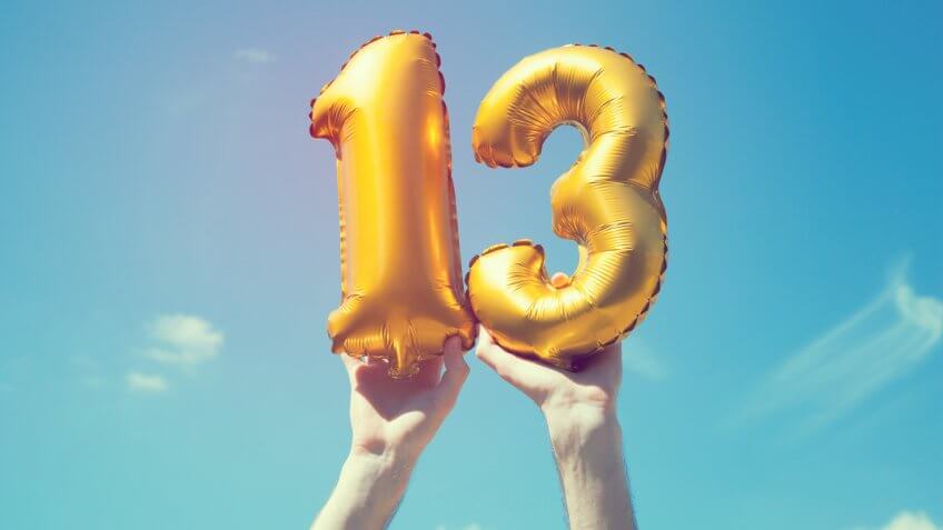 A gold foil number 13 balloon is held high in the air by caucasian male hand.