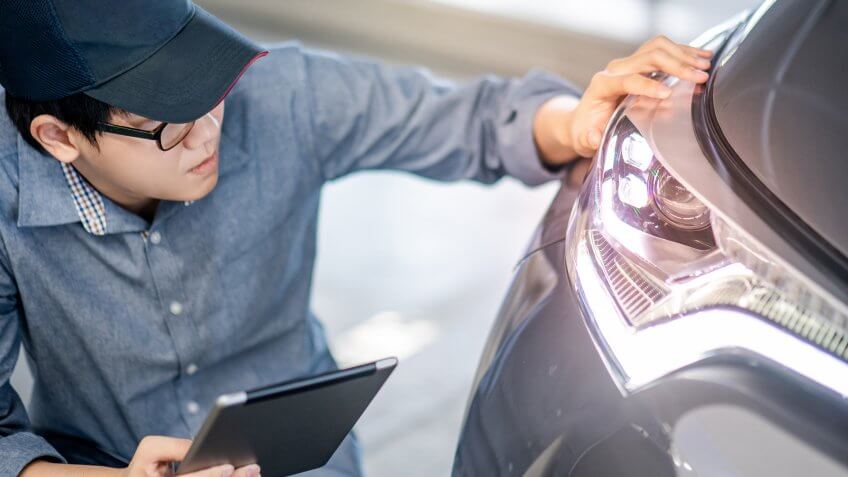 Young Asian auto mechanic holding digital tablet checking headlight in auto service garage.