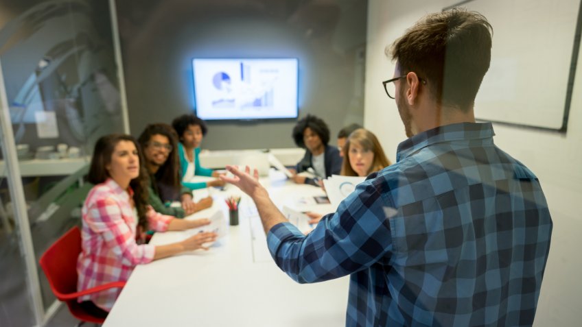 Man leading a business meeting with young people all looking at him with surprise and dissapointment with the screen on the background showing the sales statistics.