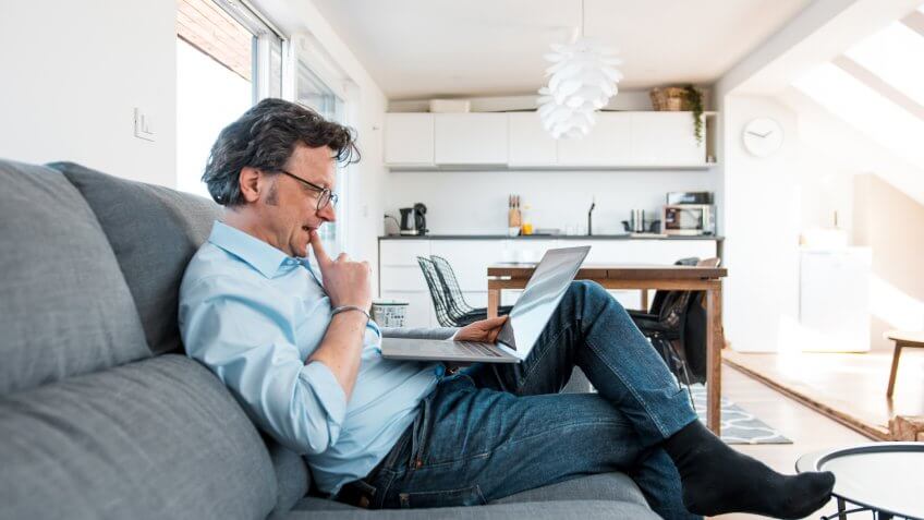 Mature Caucasian man using a laptop and sitting on the sofa in a living room.