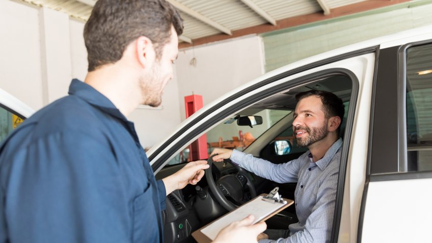 Service man holding clipboard while talking with client in car at auto repair shop.