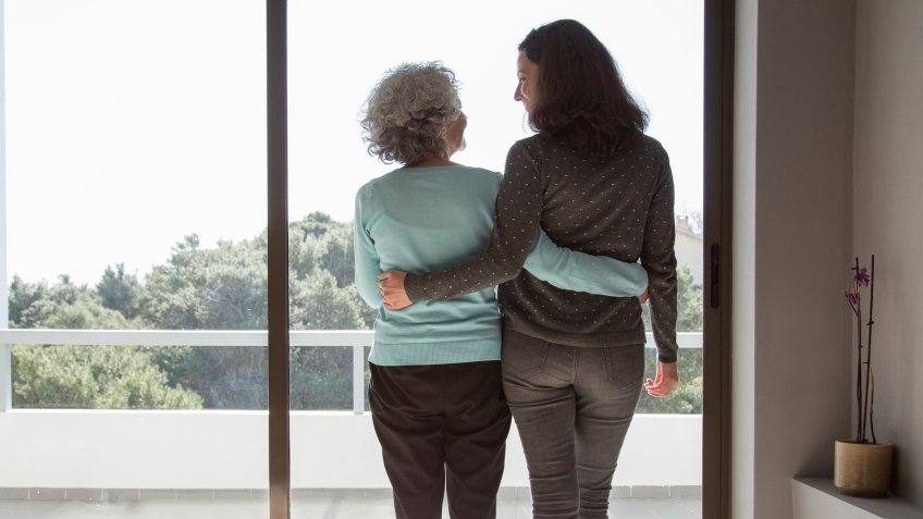 Rear view of happy mother and daughter standing embracing at window.