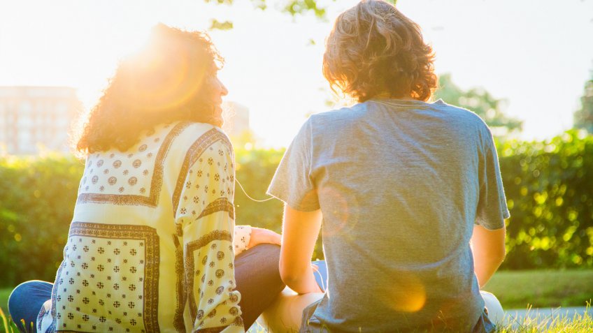 Rear view of mother sharing music sitting with teenager son in a park.
