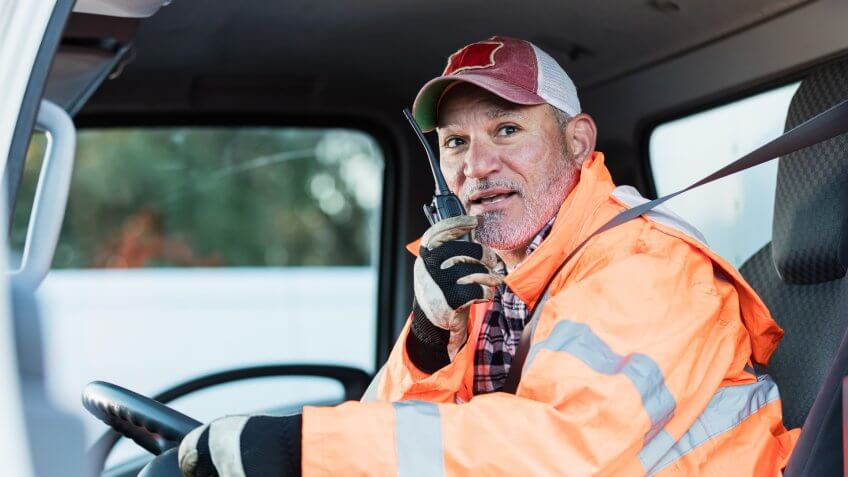 A mature Hispanic man in his 50s, a truck driver, is on his walkie-talkie, sitting in the driver's seat.