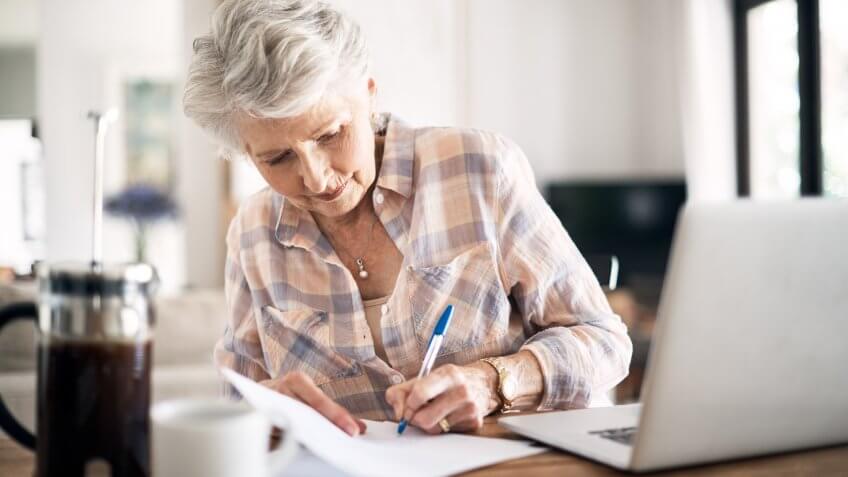 Shot of a happy senior woman flliing in paperwork while using her laptop at home.