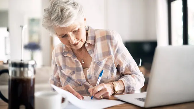 Shot of a happy senior woman flliing in paperwork while using her laptop at home.