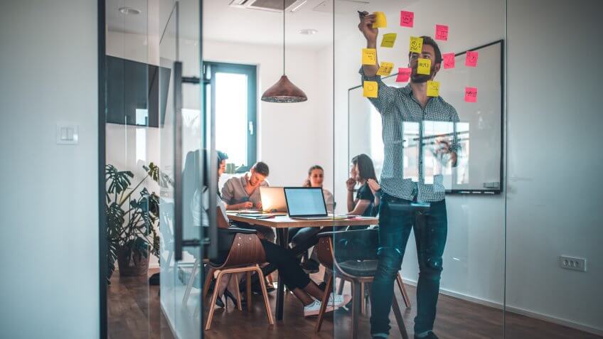 Male web designer sticking adhesive note while colleagues in background at office.