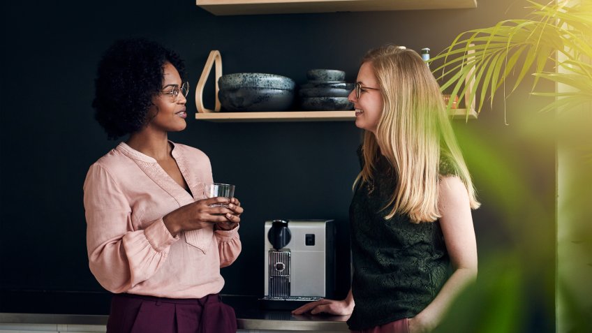 Two diverse female work colleagues smiling and talking while taking a break together in a modern office.