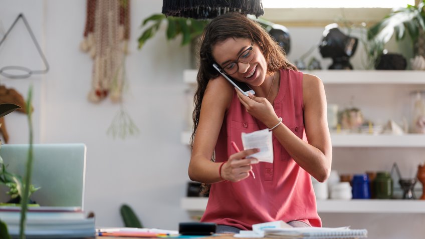 A young female sitting at a plant store holding the phone with the opposite to her ear and looking at a store receipt while smiling.