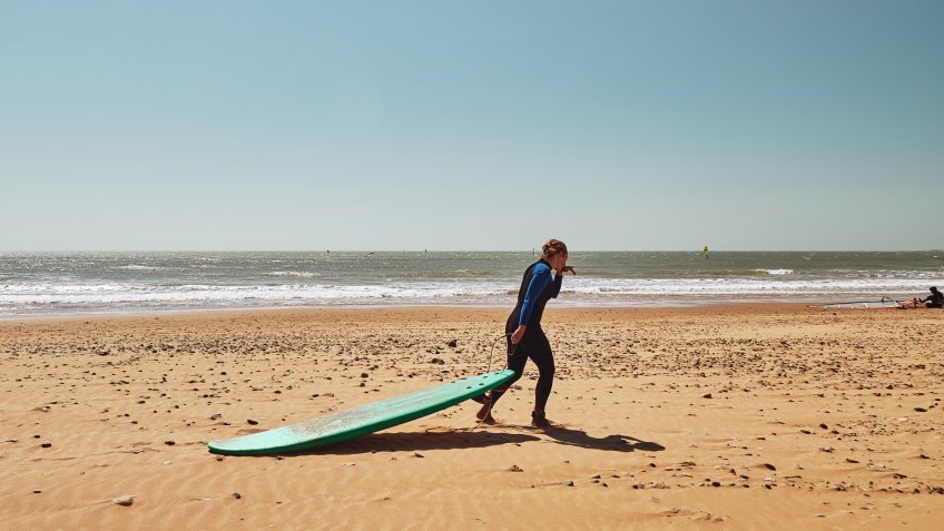 Surfer girl drags the surfboard across sand beach on a sunny day, Morocco.