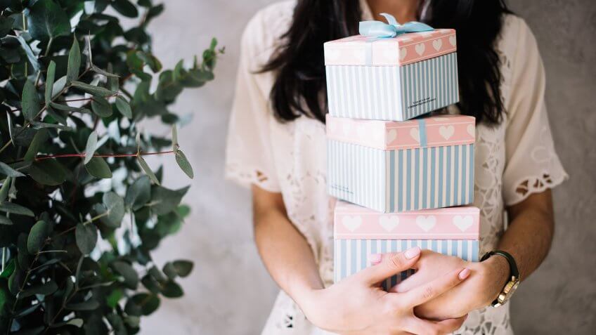 Very nice young woman holding piled colourful gift boxes with eucalyptus on the background.