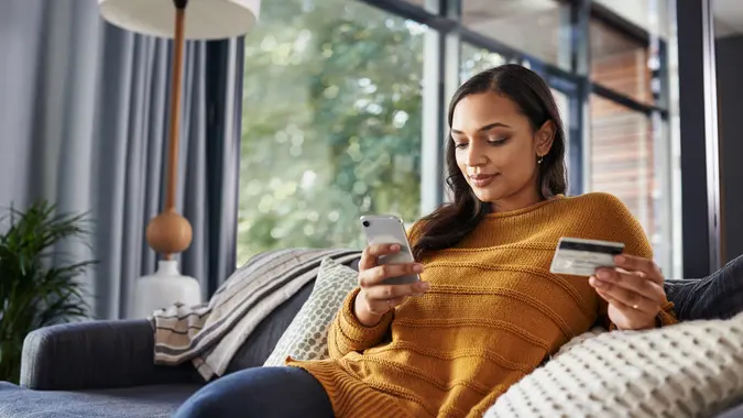 Shot of a beautiful young woman using her cellphone and credit card while relaxing on a couch at home.