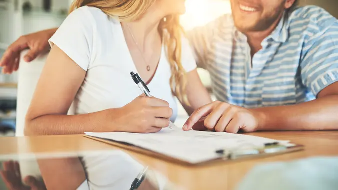 Young couple sitting together at home, smiling as they sign a document on a table, cosigning on a loan.