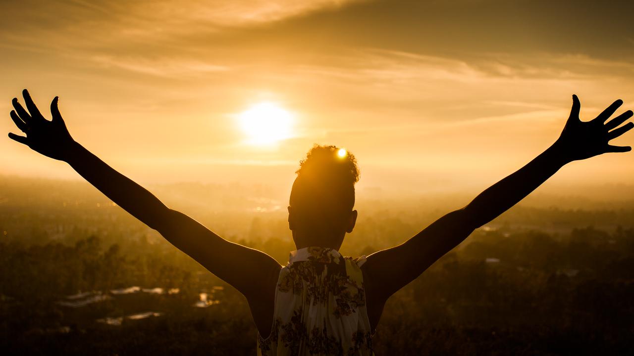 A young African American Woman raises her arms facing the sunset down over the valley and the ocean.