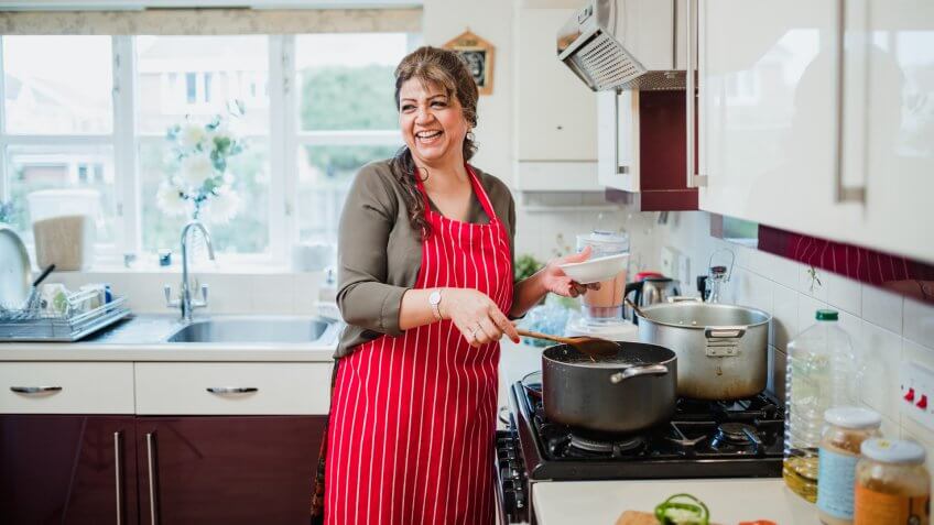 Mature woman is talking and laughing with someone out of the frame while she makes dinner at the cooker.