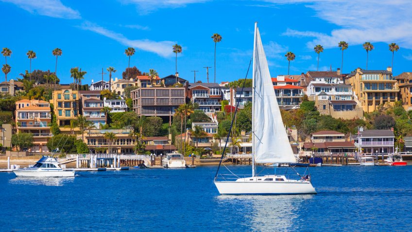 Recreational sailboat sailing on calm harbor waters of Newport Bay with beach houses and Fashion Island in background.