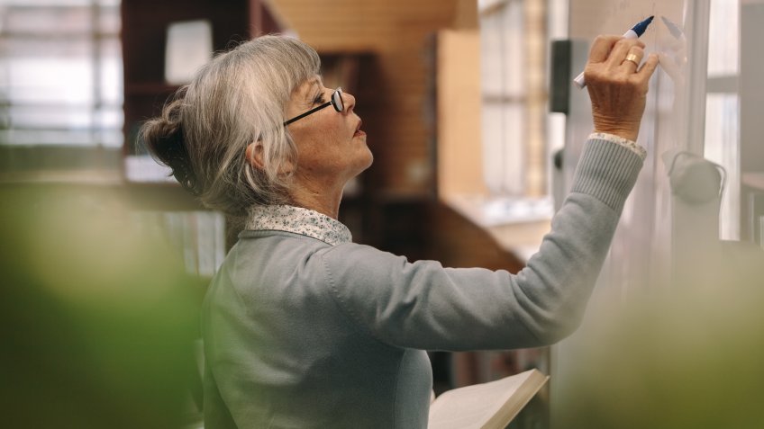 Side view of a senior female teacher writing on a white board in classroom.