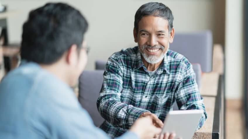Happy old short beard asian man sitting, smiling and listen to partner that showing presentation on smart digital tablet.
